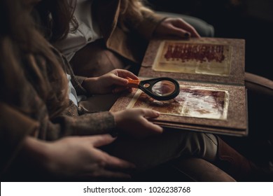 Child Girl With Woman In Image Of Detective Sits In Armchair And Looks Old Photo Album With Magnifier. Closeup.