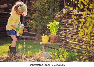 Child Girl In Yellow Cardigan Watering Flowers In Spring Garden. Outdoor Seasonal Activities For Kids. Children On The Farm, Cozy Mood, Eco Natural Lifestyle