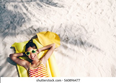 Child Girl In Yellow Swimwear And Sunglasses Chilling On Lilo Under Palm Tree On Sand At The Beach. Tropical Island. Summer Holiday Idyllic. Top View.