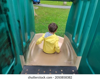 A Child Going Down A Slide From The Top. The Young Boy Is Wearing A Rain Jacket And Pushing Himself Down The Winding, Turning Slide.