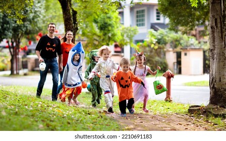 Child in Halloween costume. Asian and Caucasian kids and parents trick or treat on street. Little boy and girl with pumpkin lantern and candy bucket. Baby in witch hat. Autumn holiday fun.