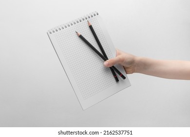 Child Hand Holding Blank Notebook With Two Pencils Above A White And Gray Background, Copy Space