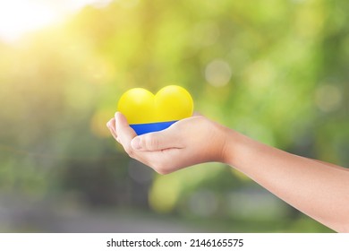 Child Hand Holding Heart In The Color Flag Nation Of Ukraine Over Green Nature Background. Concept Of Ending The War In Ukraine.