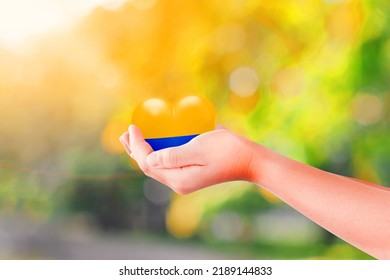 Child Hand Holding Heart In The Color Flag Nation Of Ukraine Over Green Nature Background. Concept Of Ending The War In Ukraine.