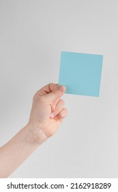 Child Hand Holding A Square Blue Blank Reminder Or Paper Notes Above A White And Gray Background, Copy Space