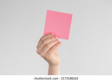 Child Hand Holding A Square Pink Blank Reminder Or Paper Notes Above A White And Gray Background, Copy Space