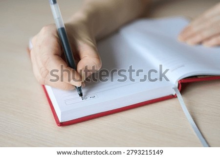Child Hand Writing Numeric Entry In Red Bound Planner On Wooden Table, Pen Held Between Fingers Over Lined Date Box, Indoor Natural Light Close-Up Showing Organizational Planning Task.