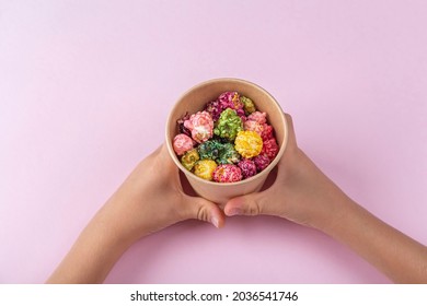 Child Hands Holding Box With Colorful Rainbow Caramel Candy Popcorn On Pink Background. Cinema Snack Concept. Watching Movie And Entertainment. Flat Lay. 