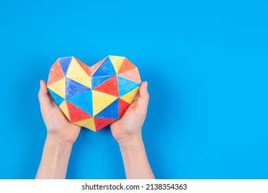Child hands holding colorful polygonal paper origami heart on light blue paper background. World autism awareness day concept