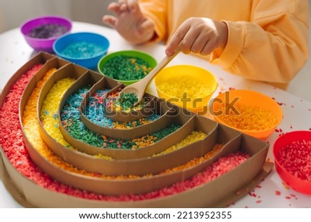 Child hands playing colored rice and make rainbow. Child filled the rainbow with bright rice. Montessori material. Sensory play and learning colors activity for kids. 