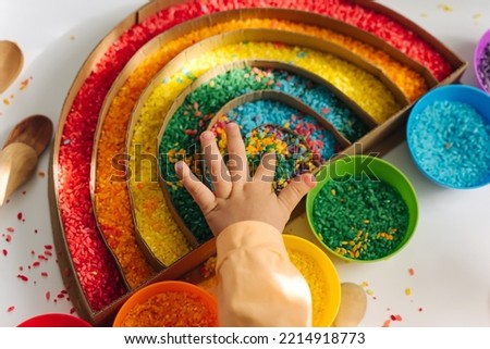 Child hands playing colored rice and make rainbow. Child filled the rainbow with bright rice. Montessori material. Sensory play and learning colors activity for kids. 