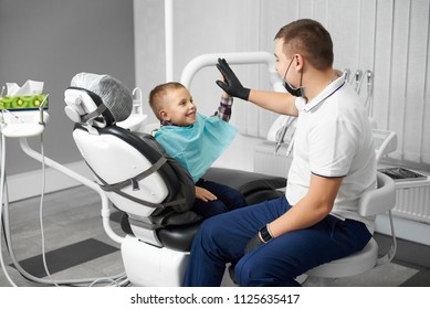 A Child Is Happy After Dental Treatment And Giving High-five To His Doctor In A Modern White Dental Clinic.