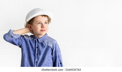 Child In A Hardhat Looks Up And Holds His Hand Behind Head. Little Boy Engineer, Architect, Builder Is Thinking, Looking For A New Idea. Choice Of Profession, Dreams Concept. Copy Space Background.