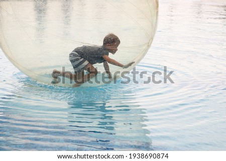 Child have fun inside big plastic balloon on the water of swimming pool on the summer resort. Little boy inside big inflatable transparent ball running and having fun. 
