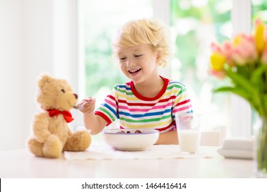 Child Having Breakfast. Kid Feeding Teddy Bear Toy, Drinking Milk And Eating Cereal With Fruit. Little Boy At White Dining Table In Kitchen At Window. Kids Eat. Healthy Nutrition For Young Kids.