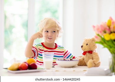 Child having breakfast. Kid feeding teddy bear toy, drinking milk and eating cereal with fruit. Little boy at white dining table in kitchen at window. Kids eat. Healthy nutrition for young kids. - Powered by Shutterstock