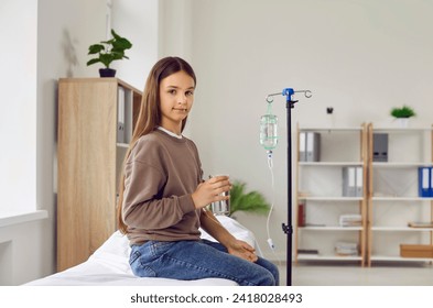 Child health care. Portrait of cute teenage girl patient in hospital with IV saline drip on her arm. Smiling Caucasian teenage girl is sitting on medical couch with glass of water and attached drip. - Powered by Shutterstock