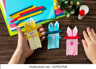 Child is holding Easter bunny toy gift stics puppets on wooden table. Handmade. Project of children's creativity, handicrafts, crafts for kids.