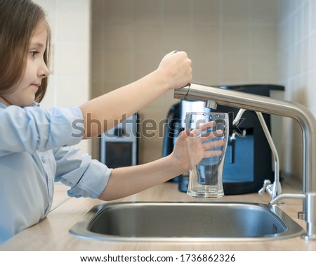 Child is holding an empty glass nearby a stainless steel kitchen tap. Person consider pouring running water from chrome faucet. Potable or Non-potable. Safe to drink or not fit for human consumption
