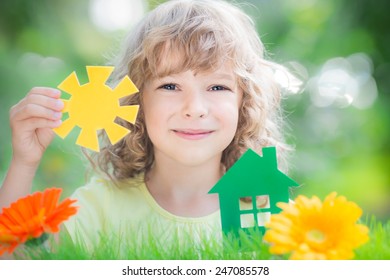 Child Holding Green Paper House And Sun In Hands Against Spring Background. Real Estate Concept