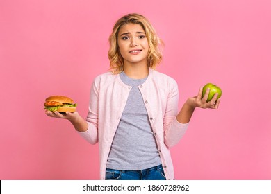 Child Holding A Piece Of Hamburger And Green Apple. Kid Girl Eats Fast Food. Not Helpful Food. Very Hungry Baby. Teen Girl Isolated Over Pink Background.