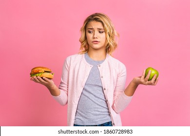 Child Holding A Piece Of Hamburger And Green Apple. Kid Girl Eats Fast Food. Not Helpful Food. Very Hungry Baby. Teen Girl Isolated Over Pink Background.