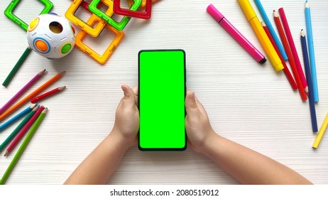 Child Holding Smartphone With Green Screen Vertical Position On White Wooden Table Background. Online Education Concept. Mobile Phone With Chroma Key. Close Up Hands.