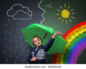 Child holding an umbrella standing in front chalk drawing changing weather from rain storm to sun shine and rainbow school blackboard