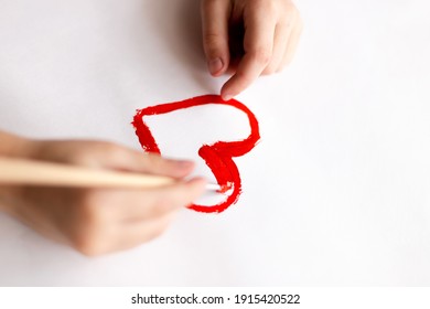 Un niño sostiene un cepillo en las manos y dibuja un corazón rojo sobre el papel blanco, vista de arriba hacia abajo. Postales para la festividad, Día de San Valentín, Día de la Madre, Día Internacional de la Mujer.