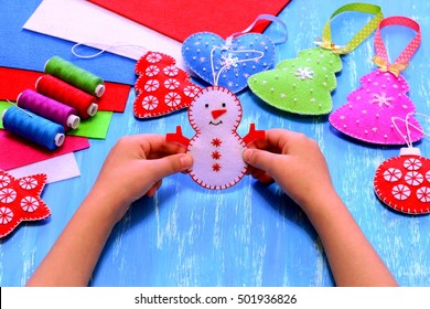 Child holds a felt snowman ornament in his hands. Christmas sewing crafts concept. Christmas tree, heart, star, snowman crafts, sewing kit, bright felt sheets on blue wooden background  - Powered by Shutterstock