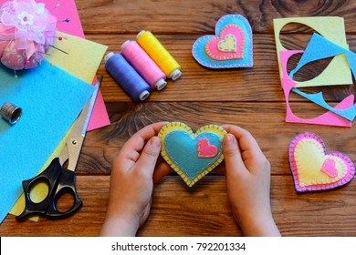 Child Holds A Felt Valentine In His Hands. Child Made Valentines From Felt. Valentines Day Crafts Idea. Felt Heart Ornaments, Scissors, Thread, Felt Sheets On A Wooden Table. Sewing Craft Projects