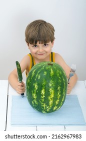 ?ute child holds a knife and fork near watermelon and looks straight. Juicy fruits or berries are very useful for children. Vitamins for babies, kids and teenagers at the white wooden table in studio.