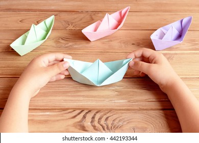 Child Holds The Origami Ship In His Hands. Colorful Ships Origami Paper Folding On A Wooden Table. Preschool And Kindergarten Paper Crafts. Summer Project Idea 