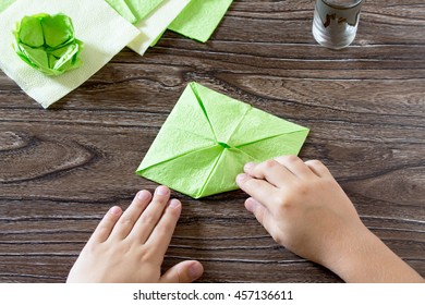 The child holds a paper square and fold it corner. The child makes crafts out of paper lily. Paper napkins on a wooden table. Children's art project, a craft for children.