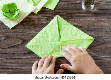 The child holds a paper square and fold it corner. The child makes crafts out of paper lily. Paper napkins on a wooden table. Children's art project, a craft for children.