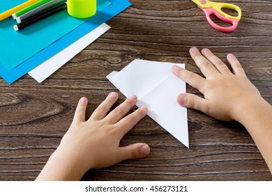 The child holds a paper strip square and fold him in half. The child makes crafts out of paper boat. Glue, paper, scissors on a wooden table. Children's art project, a craft for children.