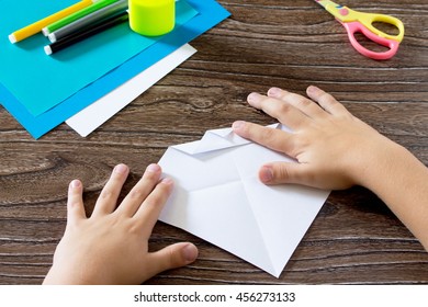 The child holds a paper strip square and fold it corner. The child makes crafts out of paper boat. Glue, paper, scissors on a wooden table. Children's art project, a craft for children.