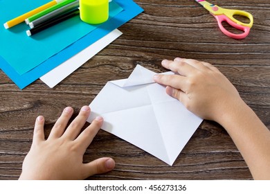 The child holds a paper strip square and fold it corner. The child makes crafts out of paper boat. Glue, paper, scissors on a wooden table. Children's art project, a craft for children.