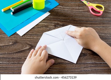 The child holds a paper strip square and fold it corner. The child makes crafts out of paper boat. Glue, paper, scissors on a wooden table. Children's art project, a craft for children.