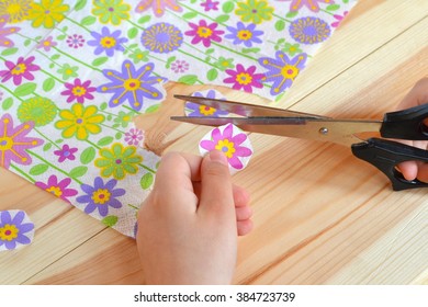 Child Holds Scissors And Paper Flower In His Hands. Child Cuts  Flowers Fragments Out Paper Napkin For Decoupage. Bright Napkin With Floral Motifs On A Wooden Table