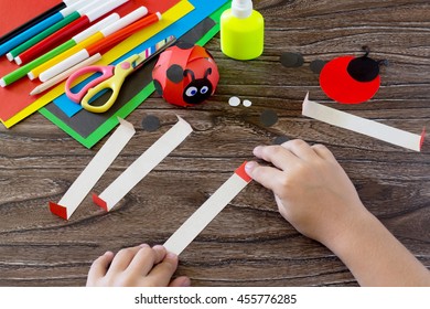 The child holds a strip of paper and fold it in half ends. The child makes crafts out of paper ladybug. Glue, paper, scissors on a wooden table. Children's art project, a craft for children.