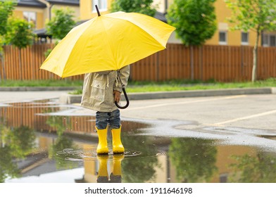 A Child Holds A Yellow Umbrella Standing In A Puddle Outside. On Sunny Day Happy Kid Walks Near The House, Plays, Has Fun