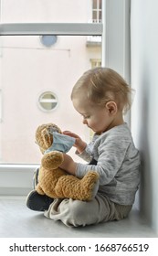 Child in home quarantine playing at the window with his sick teddy bear wearing a medical mask against viruses during coronavirus and flu outbreak. Children and illness COVID-2019 disease concept