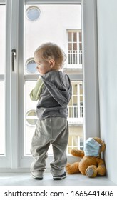 Kind in Hausquarantäne, das am Fenster steht, mit seinem kranken Teddybär, der während des Ausbruchs von Coronavirus und Grippe eine Maske gegen Viren trägt. Kinder und Krankheit COVID-2019 Krankheitskonzept