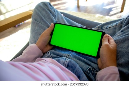 Child At Home Sits In A Chair Using A Smartphone With A Green Screen Mockup In Horizontal Landscape Mode. Using A Mobile Phone, Browsing The Internet, Watching Content, Videos, Blogging, Playing