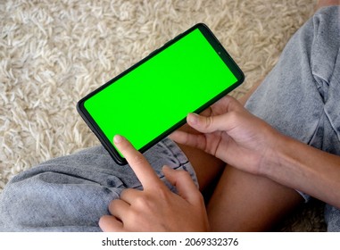 Child At Home Sits In A Chair Using A Smartphone With A Green Screen Mockup In Horizontal Landscape Mode. Using A Mobile Phone, Browsing The Internet, Watching Content, Videos, Blogging, Playing