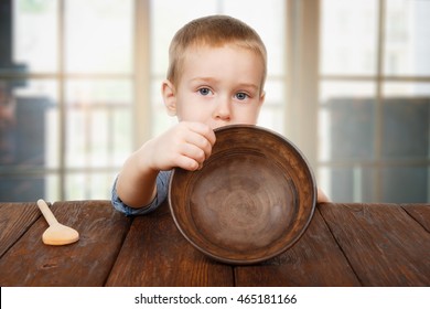 Child hunger concept. Small toddler boy shows empty bowl sitting at dark wood table with wooden spoon. Cute child has no food in plate