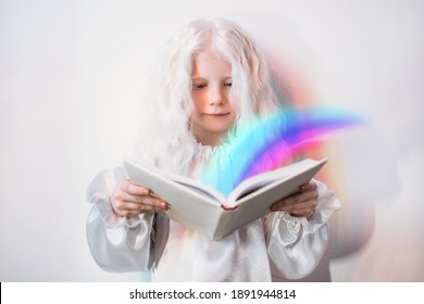 Child Imagination. Story Time. Fairytale Magic. Portrait Of Curious Sweet Blonde Little Girl In White Vintage Dress With Bright Rainbow Flowing From Open Book On Light Background.