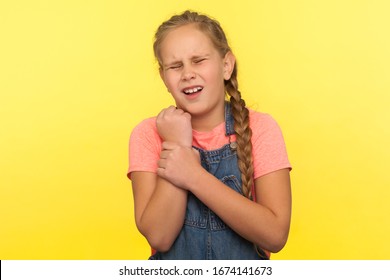 Child With Injured Hand. Portrait Of Upset Little Girl In Denim Overalls Standing With Grimace Of Pain And Touching Sore Sprained Wrist, Medical Problem. Studio Shot Isolated On Yellow Background