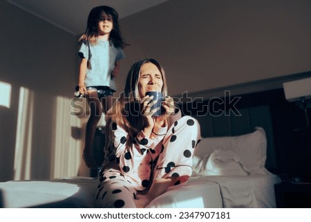 
Child Jumping in Bed Next to Exasperated Mom Drinking Coffee. Funny mother feeling overwhelmed by hyperactive kid 
